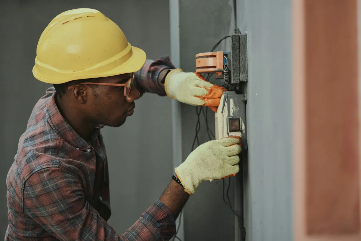 An electrician in a yellow hard hat and work gloves wiring an outlet box